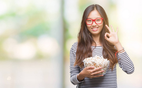 年轻的亚洲女人在孤立的背景下吃爆米花，做个好手势
