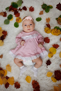 两个月大的小可爱女孩穿着一件粉红色的连衣裙，头上有一朵花，躺在被红黄秋叶包围的床上。