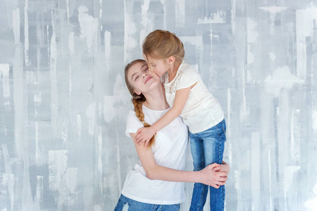 两个快乐的孩子站在灰色纹理的墙壁背景和拥抱。可爱的漂亮小女孩拥抱紧身可爱的少女，表现出她的爱和关怀。姐妹们在家玩得开心