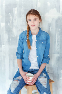年轻快乐可爱的十几岁女孩穿着牛仔裤，牛仔夹克和白色T恤坐在椅子上，灰色纹理的墙壁背景，拿着纸杯喝咖啡。休闲日常生活方式