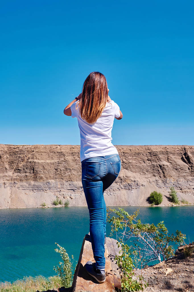 一个年轻漂亮的女孩站在蓝色湖面上的悬崖上,透过望远镜眺望远方