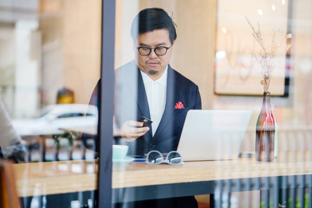 一个穿着马球和夹克的中国人白天坐在咖啡馆里用笔记本电脑工作的肖像。 他在网上购物