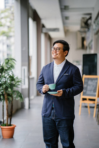 一位受过教育的中国中年人的肖像，穿着商务休闲和眼镜。 他白天在城里拿着一个优雅的杯子和茶碟喝茶。