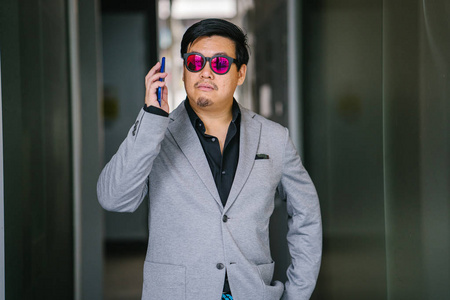 一位中年中国亚裔新加坡人男子的肖像，他穿着黑色衬衫灰色西装和一副时尚的反光太阳镜。 他在用智能手机