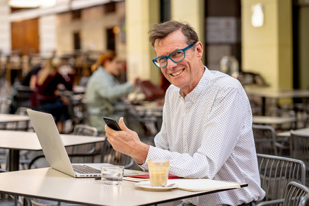 微笑老人检查智能手机，同时在露台户外咖啡店的电脑上工作，老年人使用现代技术保持联系和企业家创意商业理念。