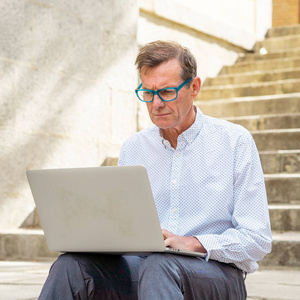时尚的老人在笔记本电脑上工作，上网，坐在户外城市的楼梯上，在数字游牧中，老年人使用现代技术保持联系和企业家创意的商业理念。