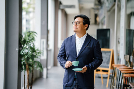 一位受过教育的中国中年人的肖像，穿着商务休闲和眼镜。 他白天在城里拿着一个优雅的杯子和茶碟喝茶。