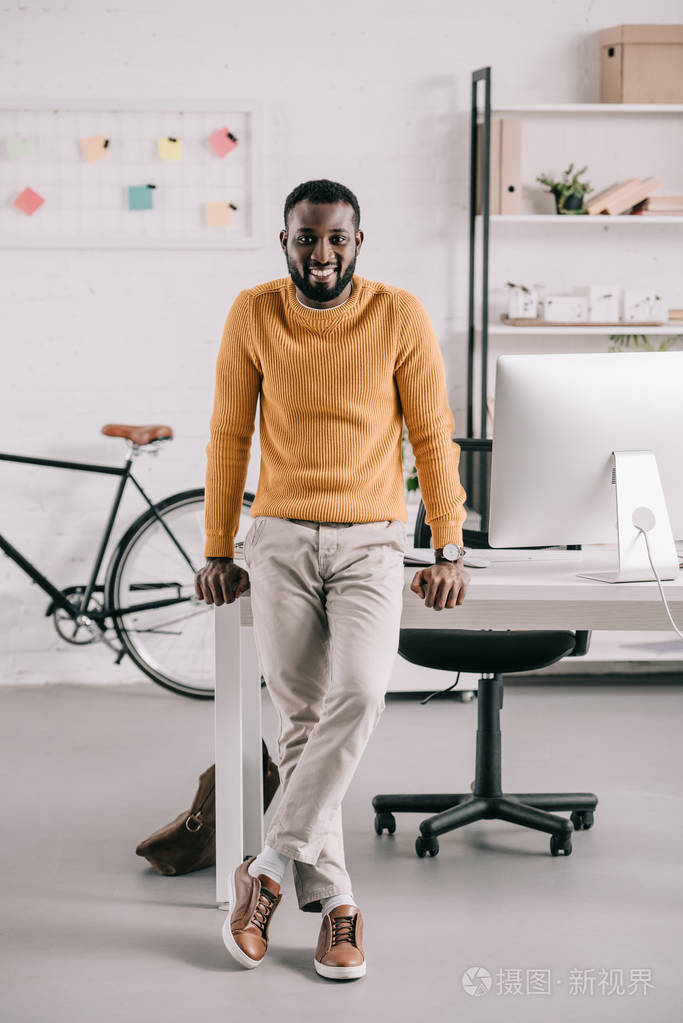 微笑着,英俊的非裔美国设计师,穿着橙色毛衣,靠在桌子上,在办公室里