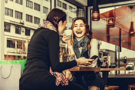 两个漂亮的微笑女孩在购物后在咖啡馆喝咖啡时使用智能手机。 友谊和技术。