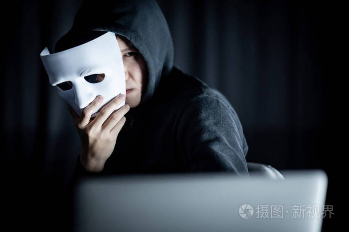 神秘男帽衫黑客拿着白色面具看着笔记本电脑匿名社交伪装网络攻击或