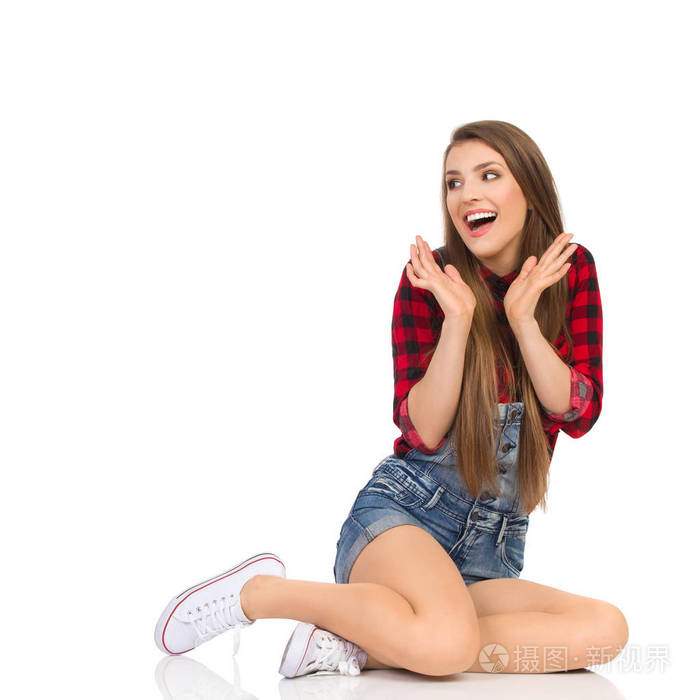 美丽的年轻女子穿着红色伐木工人衬衫牛仔裤短裤和白色运动鞋坐在地板