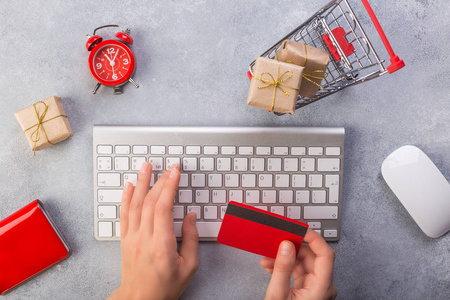 女人用信用卡在线订购礼物。 概念网上购物购买礼物。 红色信用卡Keyborad和圣诞礼物在灰色桌子上平躺复制空间。