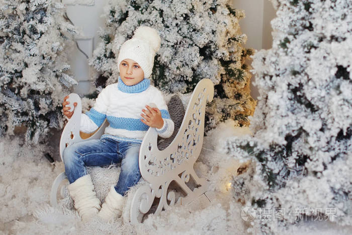 穿着白色和蓝色毛衣和白色帽子的男孩坐在圣诞树附近的装饰雪橇上。