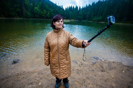 幸福的老年妇女欣赏美丽的大山湖景色。森林湖附近的高级女游客在动作摄像机上自拍，和谐理念