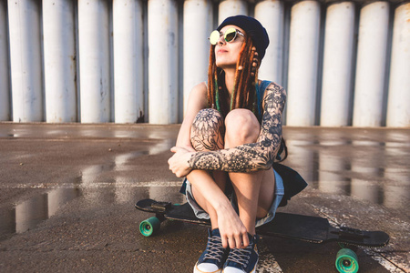与纹身和长发绺在蓝色帽子的年轻女孩坐在长板的背景下混凝土结构