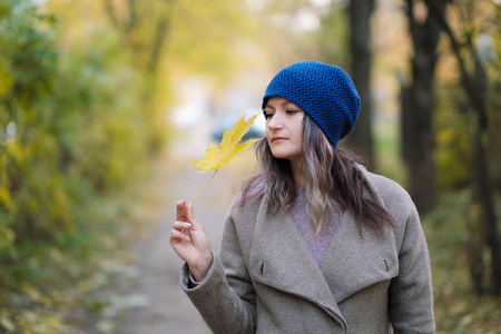 在秋天的树木和枫叶的背景的大衣和蓝色帽子的女孩