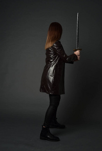 穿红色长皮杰克的黑发女孩的全长肖像。 站立姿势，手持剑在灰色工作室背景。