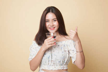 年轻的亚洲女人竖起大拇指，用米色背景的一杯饮用水