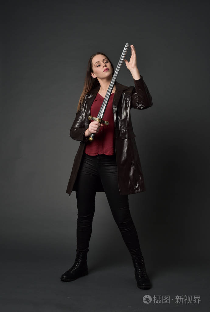 穿红色长皮杰克的黑发女孩的全长肖像站立姿势手持剑在灰色工作室背景
