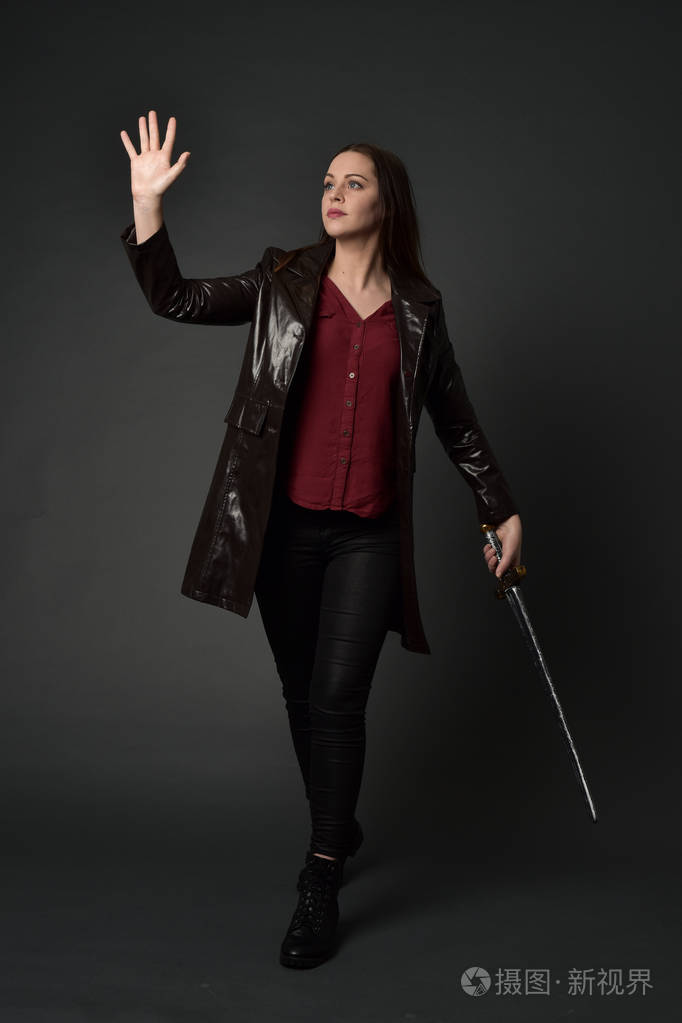 穿红色长皮杰克的黑发女孩的全长肖像站立姿势手持剑在灰色工作室背景