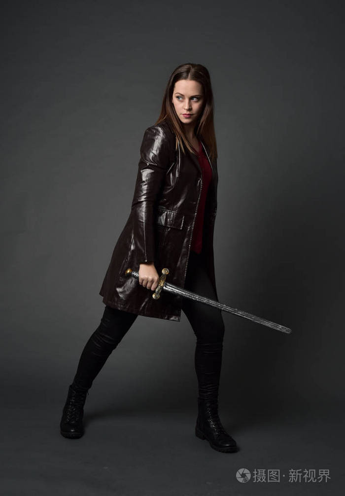 穿红色长皮杰克的黑发女孩的全长肖像站立姿势手持剑在灰色工作室背景
