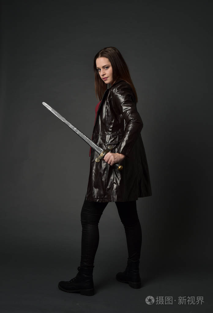 穿红色长皮杰克的黑发女孩的全长肖像站立姿势手持剑在灰色工作室背景