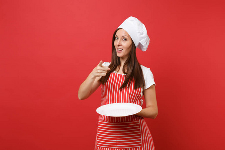 家庭主妇女厨师或面包师穿条纹围裙，白色T恤，烤厨师帽子隔离在红墙背景上。 女人拿着空的空白圆板和食物的地方。 模拟复制空间概念