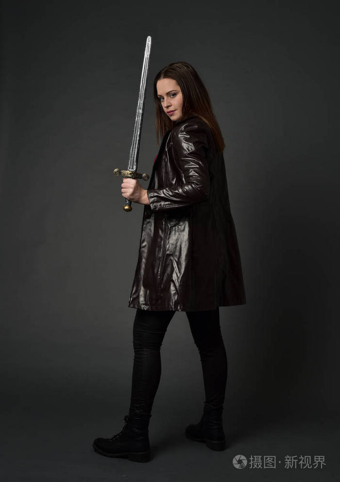 穿红色长皮杰克的黑发女孩的全长肖像站立姿势手持剑在灰色工作室背景