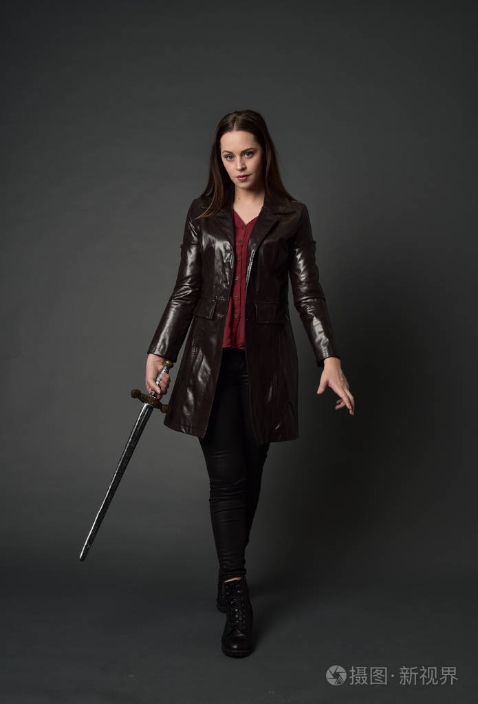 穿红色长皮杰克的黑发女孩的全长肖像站立姿势手持剑在灰色工作室背景