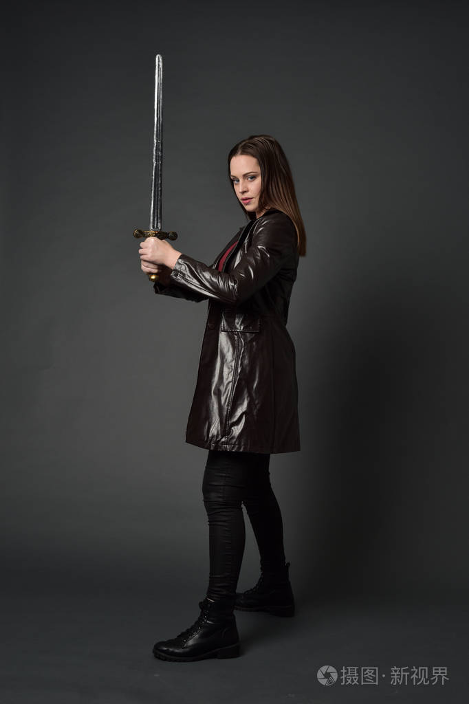 穿红色长皮杰克的黑发女孩的全长肖像站立姿势手持剑在灰色工作室背景