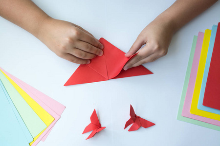 s hands do origami butterfly from colored paper on white backgro