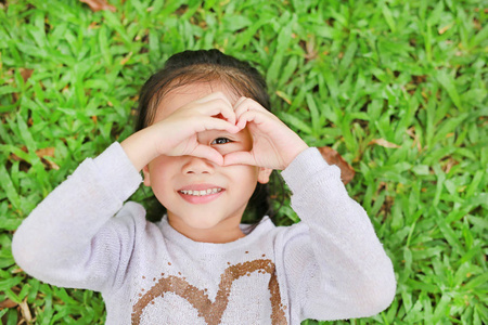 微笑可爱的亚洲小女孩躺在绿色草坪上的肖像,让她的手在眼睛上做心脏