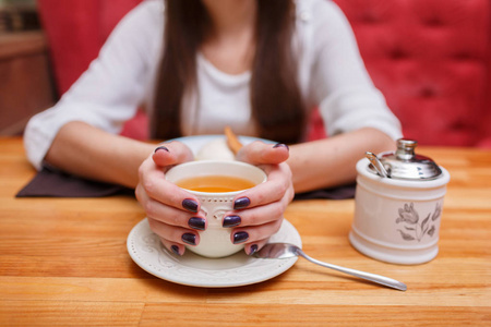 靠近女性的手，用深色的指甲拿着一杯茶和一罐糖，靠近咖啡馆模糊的背景。 热饮概念