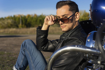 穿着黑色皮夹克太阳镜和牛仔裤的年轻人坐在户外的摩托车上，在蓝天的背景下。 生活方式旅行。 复制空间。 广告和商业设计。