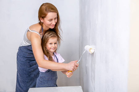 在公寓里修理。 幸福的家庭母女穿着围裙用白色油漆粉刷墙壁。 母亲帮助女儿用滚筒粉刷墙壁。 水平射击