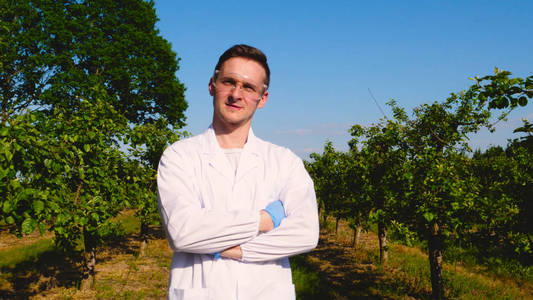 一位年轻英俊的男性生物学家或农学家，穿着白色外套，戴着蓝色橡胶手套，穿过苹果树，了解大自然和绿色植物的背景。