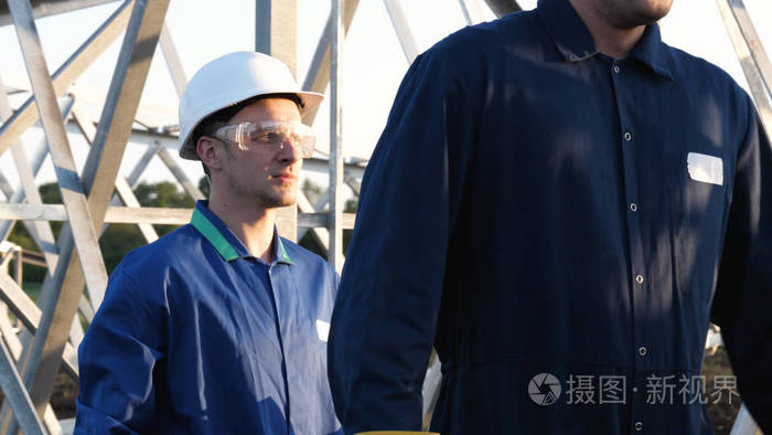 电工或建筑工人和工程师穿着蓝色长袍，戴着黄色和白色头盔，检查技术概述，以了解强度，准备电线杆铁塔能源和工人。