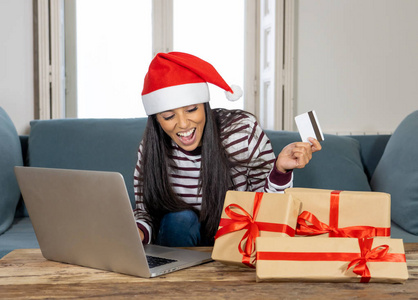 穿着红色毛衣戴着圣诞帽拿着信用卡在家中使用笔记本电脑购买圣诞礼物的妇女兴奋不已，网上购物和信用卡设施令人兴奋