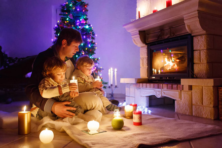 父亲和两个蹒跚学步的小男孩坐在烟囱, 蜡烛和壁炉, 并期待着火。家庭庆祝圣诞节。有圣诞树和灯光的背景。孩子们喜欢礼物