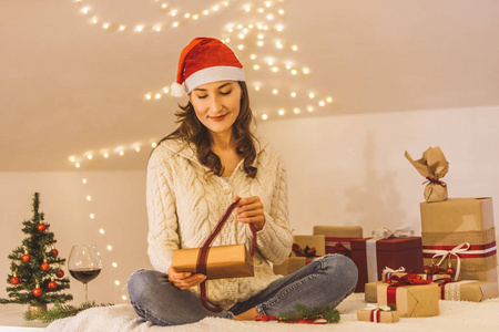 年轻美丽的微笑女人为她的朋友和家人包装包装或包装圣诞礼物，戴上红色圣诞老人帽，准备圣诞节假期和庆祝概念