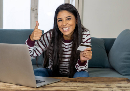 吸引人的年轻拉丁女性持有信用卡，并使用笔记本电脑在家里在互联网上购买在线商务和技术数字营销休闲生活方式的概念。