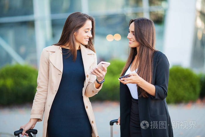 两个快乐的欧洲女性看着智能手机，同时拿着行李站在机场附近等待航班或起飞后。空中旅行概念。