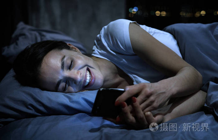 约会app概念.年轻女子晚上躺在床上,看着智能手机,笑着发短信