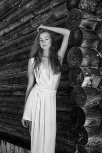 的年轻女人穿着一件浅白色的连衣裙在老房子附近的村庄里摆姿势。 在一个圆木的乡间别墅的背景上有一个完美身材的女孩