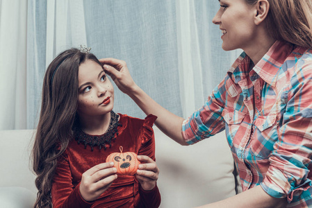 微笑的女人和穿着服装的小女孩在沙发上。 美丽的母亲帮助可爱的微笑女儿参加万圣节派对。 庆祝万圣节。 家庭和儿童概念