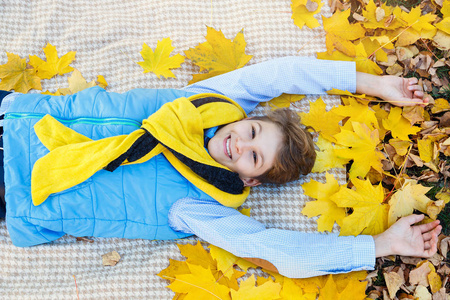可爱的男孩穿着蓝色衬衫微笑，黄色的围巾躺在米色格子上，上面有枫叶。秋天心情美好的一天。平静生活中的黄金坠落。明亮的坠落图像。
