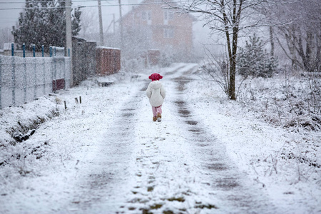 穿着灰色针织外套的小女孩走在冬天的雪道上，背景是晴朗的森林