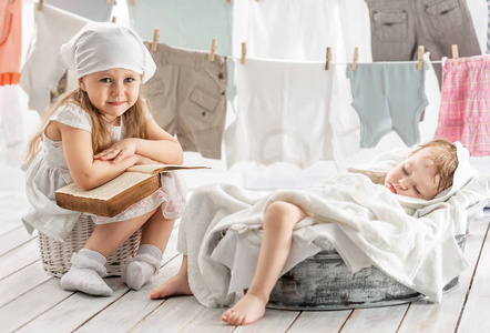 小女孩读一本书给他妹妹，她睡在碗里，洗衣房里洗衣服