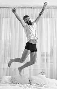 男子在衬衫和内裤跳跃在床上, 白色窗帘的背景。人在愉快的面孔充满能量在早晨。有胡子的男人在空中跳得很高。充满力量和能量的概念