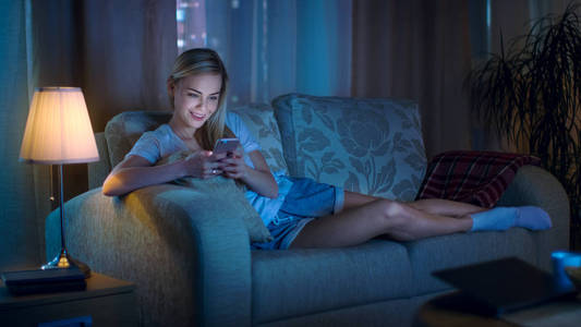 深夜,美丽的女人躺在客厅的沙发上,用她的智能手机和微笑照片